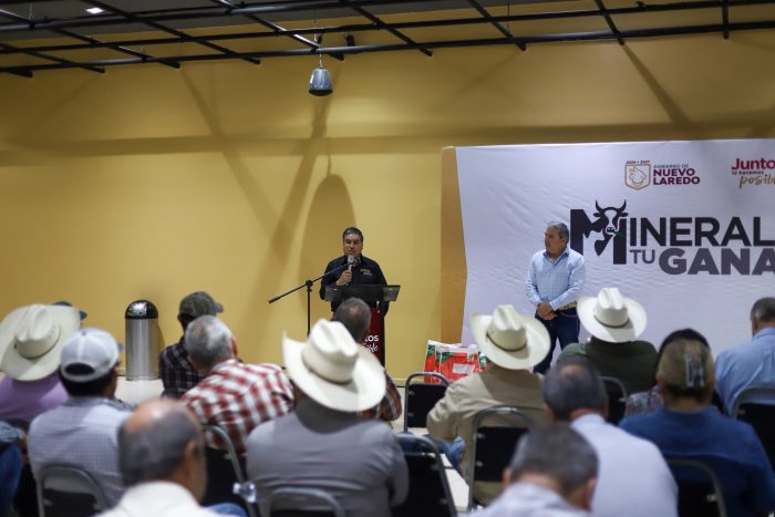 Man at a podium addressing an audience in a room; many attendees wear cowboy hats, with a stage banner reading 'MINERA TU GANA'.