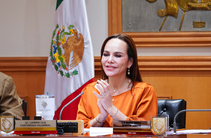 Professional woman in an orange blouse sits at a conference table, hands clasped, with a microphone in front and the Mexican flag and wood panel backdrop behind.