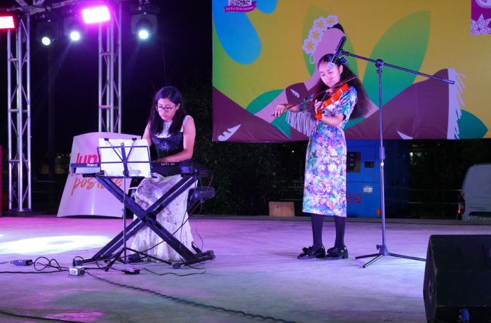 Two young girls perform on a stage at night: one playing a keyboard, the other playing a violin, under colorful backdrop and stage lights.