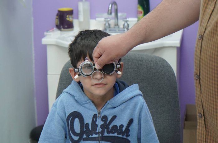 Boy in a blue hoodie sits in an exam chair while a clinician adjusts trial frames on his face in a clinic with a purple wall behind him.