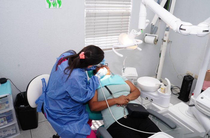 Dental hygienist in a blue gown treats a patient reclining in a dental chair with overhead light and instruments nearby.