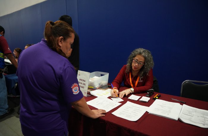 Staff member with an orange lanyard signs forms at a help desk while a visitor in a purple shirt waits.