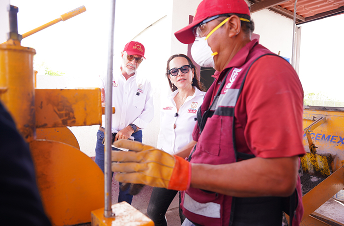 Worker in a red shirt and gloves operates orange construction equipment as two observers in hard hats and safety gear watch nearby.