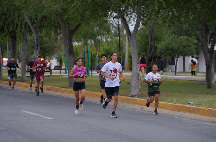 Group of runners racing down a tree-lined street during a community race, bib numbers visible on their chests.