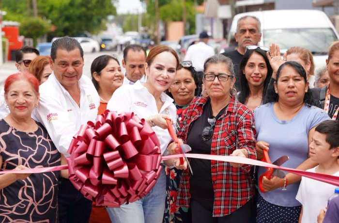 Woman in a white shirt cuts a pink ribbon at a community ribbon-cutting ceremony, surrounded by diverse smiling onlookers.