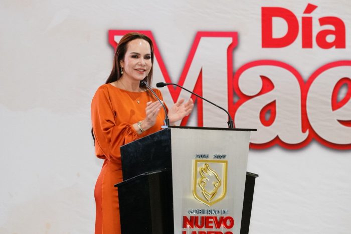 Woman in an orange dress speaks at a podium with a gold emblem, in front of a large red banner and microphone setup at a formal event.