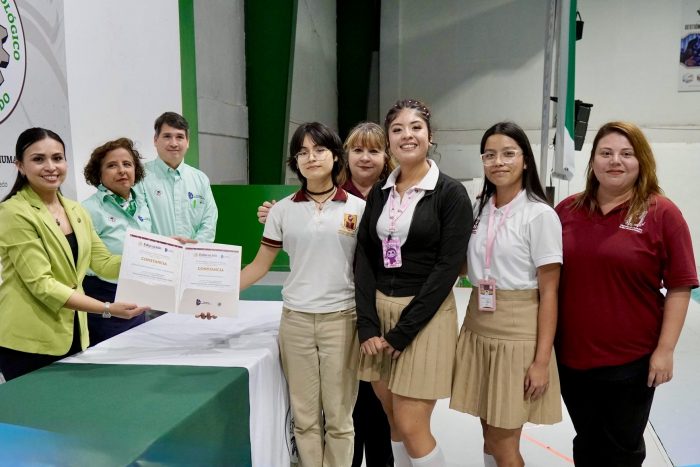 Group of students and staff pose for a certificate presentation around a table in a bright room; two people hand a document to a student in a beige uniform.