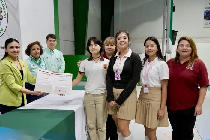 Group of students and staff pose for a certificate presentation around a table in a bright room; two people hand a document to a student in a beige uniform.