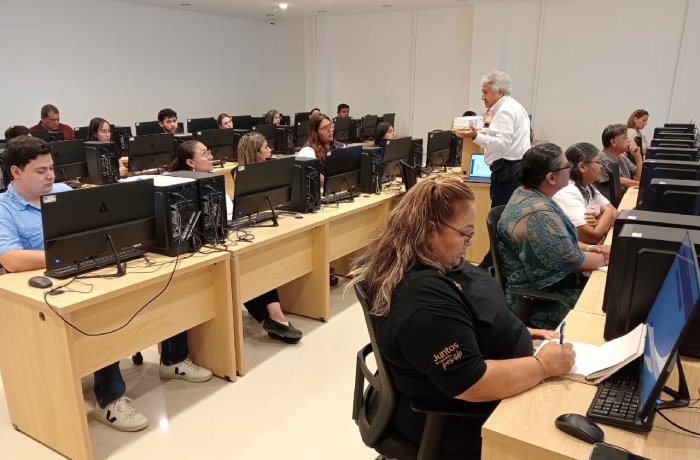 Instructor guides adult students in a computer lab, standing at the front while learners work at row after row of desks with desktop computers.
