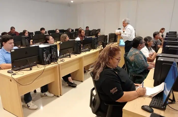 Instructor guides adult students in a computer lab, standing at the front while learners work at row after row of desks with desktop computers.