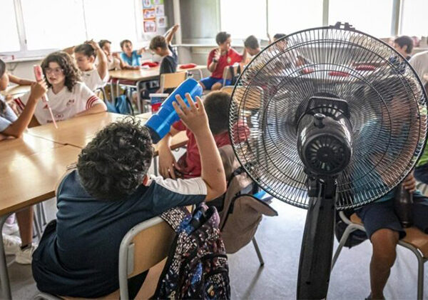 In a busy classroom, students sit at desks while a large pedestal fan blows air in the foreground.