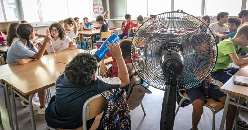 In a busy classroom, students sit at desks while a large pedestal fan blows air in the foreground.