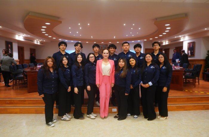 Group of students in navy shirts posing with a woman in a pink suit in a formal conference room or auditorium setting.