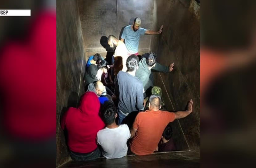 Crowd of people packed into a narrow vertical shaft, some touching the walls as they await rescue, with one man at the top supervising.