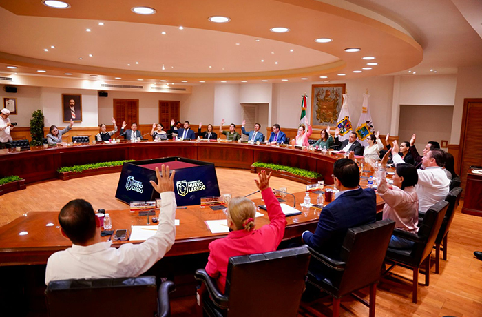 A large conference room with a circular wooden table, many participants around it, several raising their hands in a formal meeting.