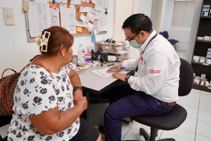 Healthcare professional wearing a mask consults with an elderly woman at a clinic desk and papers nearby.
