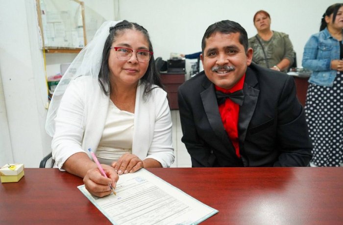 A bride in a white dress and veil signs a document on a wooden table beside a man in a black suit with a red bow tie, as others stand in the background in a formal setting.