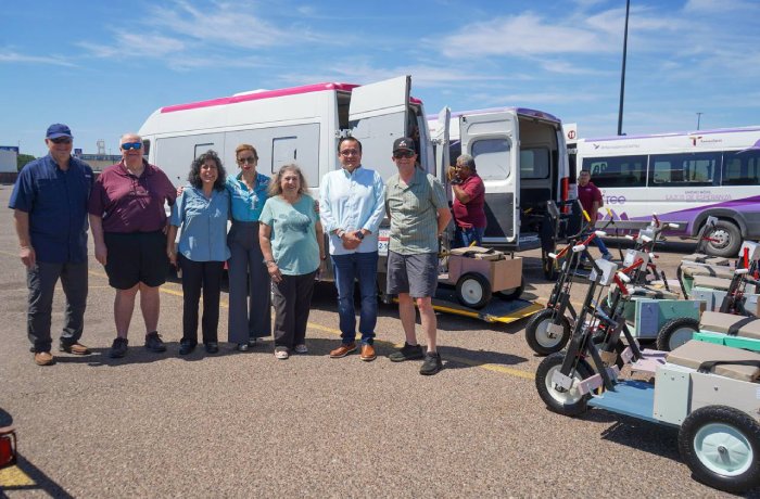 Group of nine people posing in a parking lot beside vans, with mobility scooters and equipment on the right.