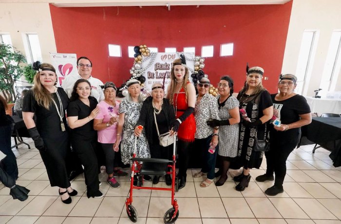 Diverse group of adults posing for a photo at a community event, with a woman in red and a walker at center and a balloon arch in back.