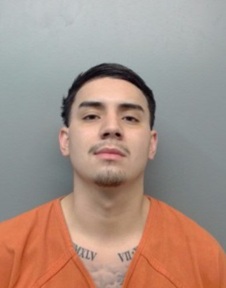 Young man with short dark hair in an orange jail uniform against a gray wall, with visible chest tattoos.