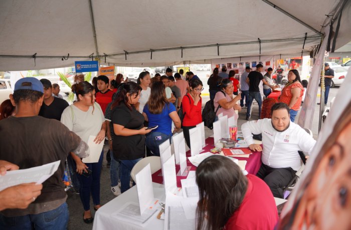 People line up under a tent at a community fair; volunteers sit at tables with papers and forms to help attendees.