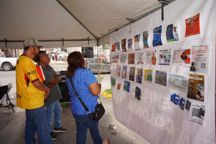 People stand under a white tent, looking at a wall of posters and flyers displayed for a community event.