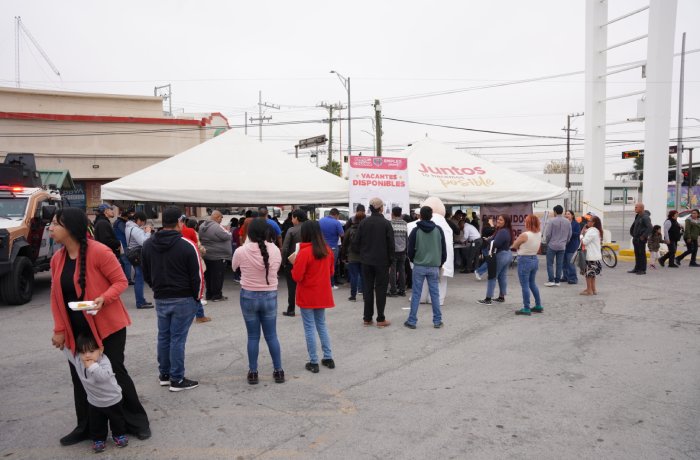 People stand in a line under a large white tent in a parking lot at a community event, likely receiving services or meals.