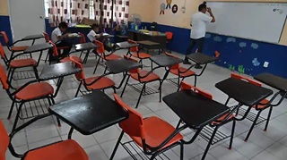 A classroom with rows of red and black student desks; a teacher stands at the whiteboard at the front of the room.