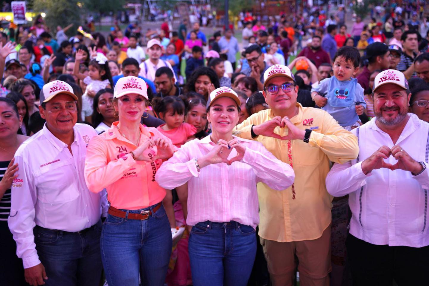 Group of people in a crowd forming heart shapes with their hands, smiling at the camera during an outdoor event.