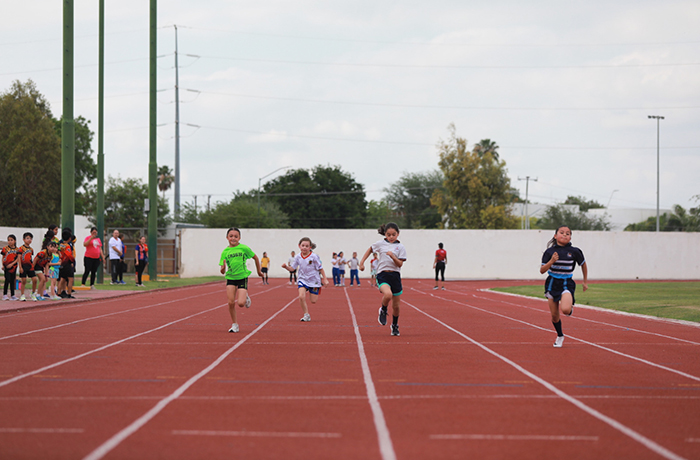 Children sprint on a red track during a race, with onlookers and a white wall in the background.