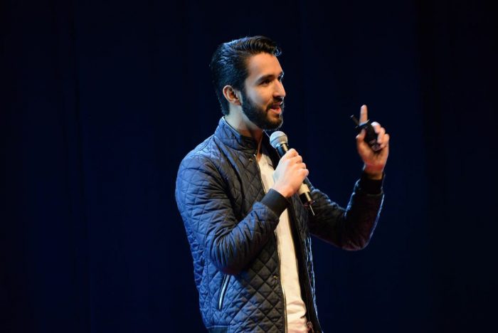 Male presenter on a dark stage, speaking into a handheld microphone and gesturing with a clicker.