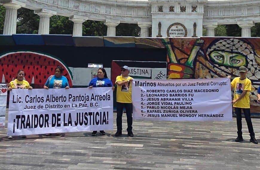 Group of protesters holding large banners with names and messages in Spanish in a plaza, in front of a colorful mural and stone colonnade.