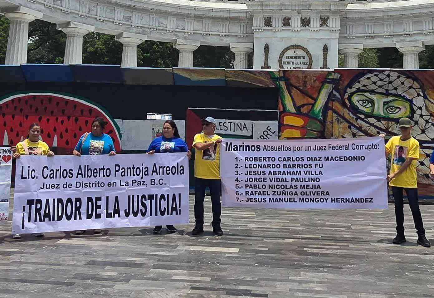 Group of protesters holding large banners with names and messages in Spanish in a plaza, in front of a colorful mural and stone colonnade.