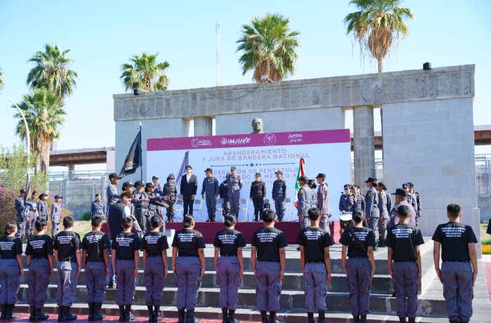 Outdoor ceremony with uniformed officers on a stage and rows of cadets in black shirts facing them; a purple banner and flags behind, with palm trees in the background.