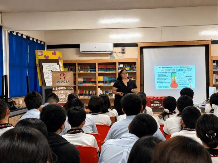 Instructor speaks to a class in a library; projector screen shows a mental health poster about suicide prevention.