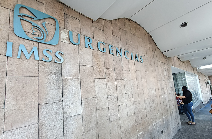 Hospital entrance wall with blue 'URGENCIAS IMSS' sign and logo; people near a doorway on the right.
