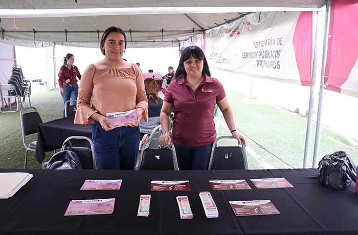 Two women stand behind a black table filled with pink brochures under a white canopy at a public service event.