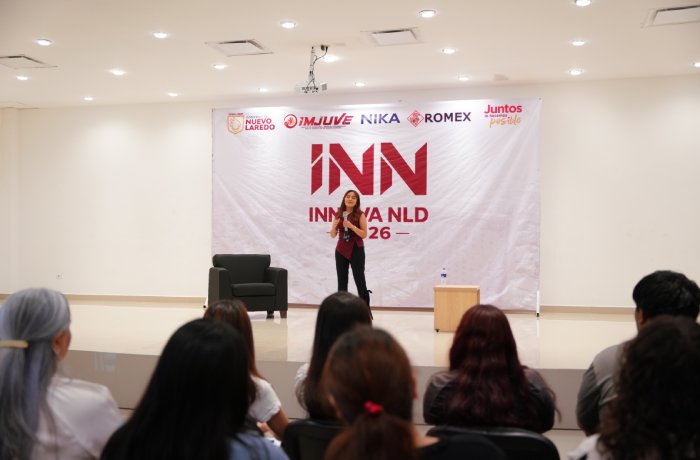 Speaker presenting on a stage in a bright hall, addressing an audience seated in front; large banner with 'INN' and logos behind her