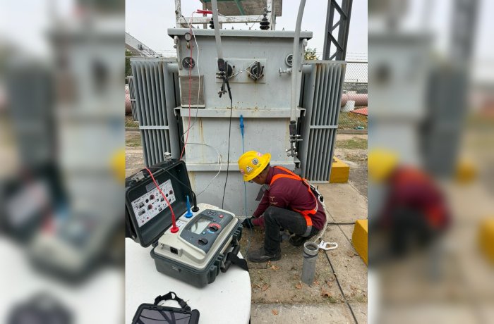 Technician in a yellow hard hat and orange safety vest kneels beside a large electrical transformer, connecting a portable testing device with red and blue leads.