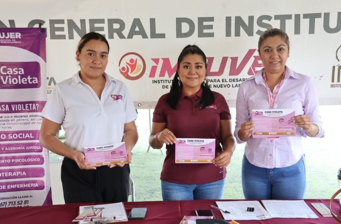 Three women stand behind a red table displaying pink informational brochures at an outreach event, with a banner and posters in the background promoting a women’s health project (Casa Violeta).