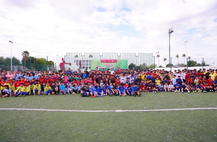 Large group of children in colorful uniforms posing for a group photo on a grassy sports field, with a banner and fences in the background.
