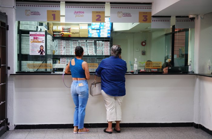 Two women stand at a service counter in a government/postal office, facing windows labeled 1–3 with clerks behind glass and shelves of boxes overhead.