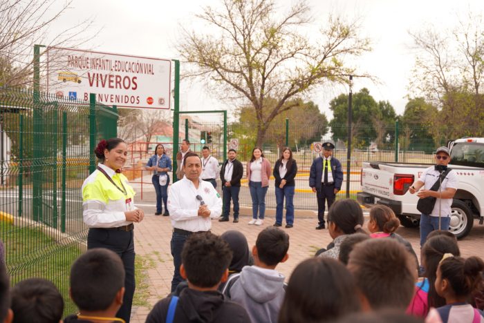 Group of adults and kids in a schoolyard during a road-safety education event; a woman in a high-visibility vest speaks to students while others stand nearby.