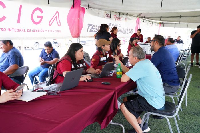 Staff in red shirts assist visitors at an outdoor help desk with laptops under a canopy/tent.
