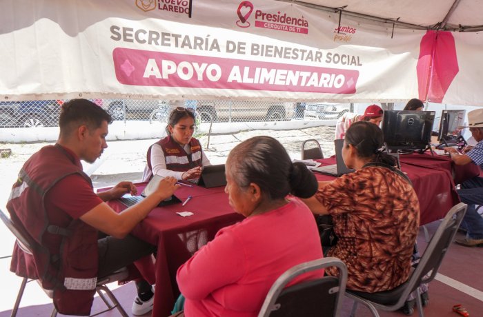 Temporary aid booth under a white tent labeled 'Apoyo Alimentario' where staff assist seated community members with documents and forms.