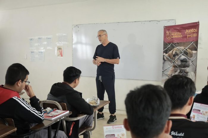 Instructor stands at the front of a classroom delivering a lesson to students seated at desks, with a whiteboard behind him and a dog poster on the right.