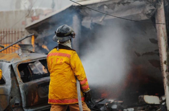 Firefighter in bright yellow turnout gear and helmet stands amid smoke at a charred vehicle fire, surveying the scene.