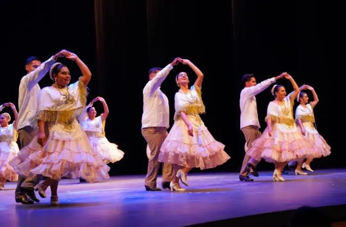 Group of dancers performing a coordinated routine on stage; women in pink ruffled dresses and men in white shirts.