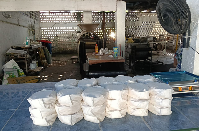 Row of white sacks lined up on a blue counter in a workshop, with a cooking area, scale, and bottles visible in the background.