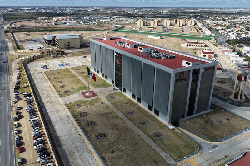 Aerial view of a large gray customs building with a red roof, labeled 'Aduanas', surrounded by parking lots and roads in an urban area.